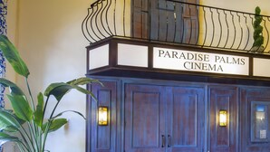 Der Eingang eines Kinos mit einem Schild, auf dem "Paradise Palms Cinema" steht. Es gibt zwei große Holztüren mit Leuchten an jeder Seite. Im Vordergrund steht ein blauer Blumentopf, und vor den Türen verläuft eine Metallbarriere.
