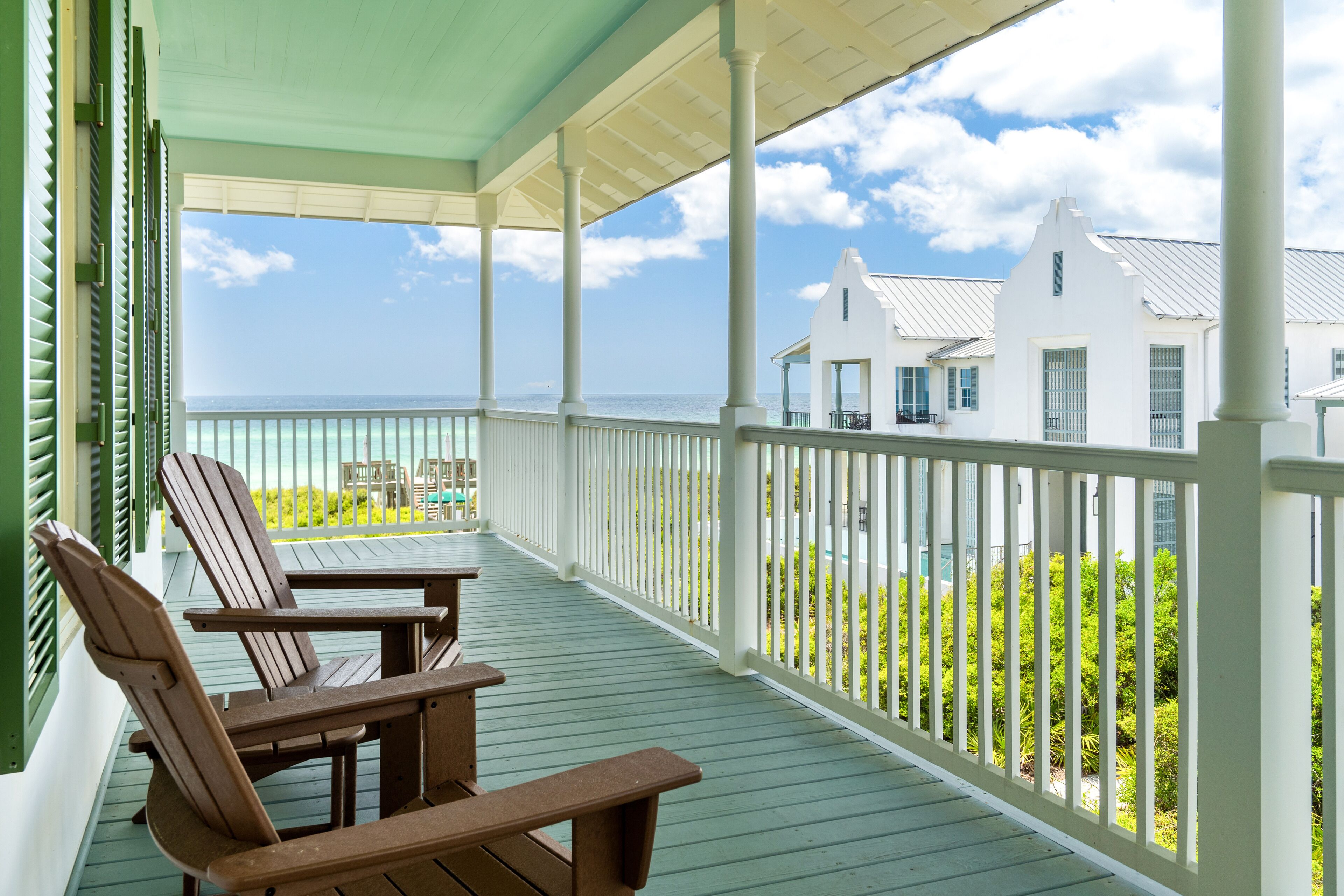 Eine überdachte Veranda mit Holzstühlen und Blick auf das Meer sowie weiße Gebäude.
