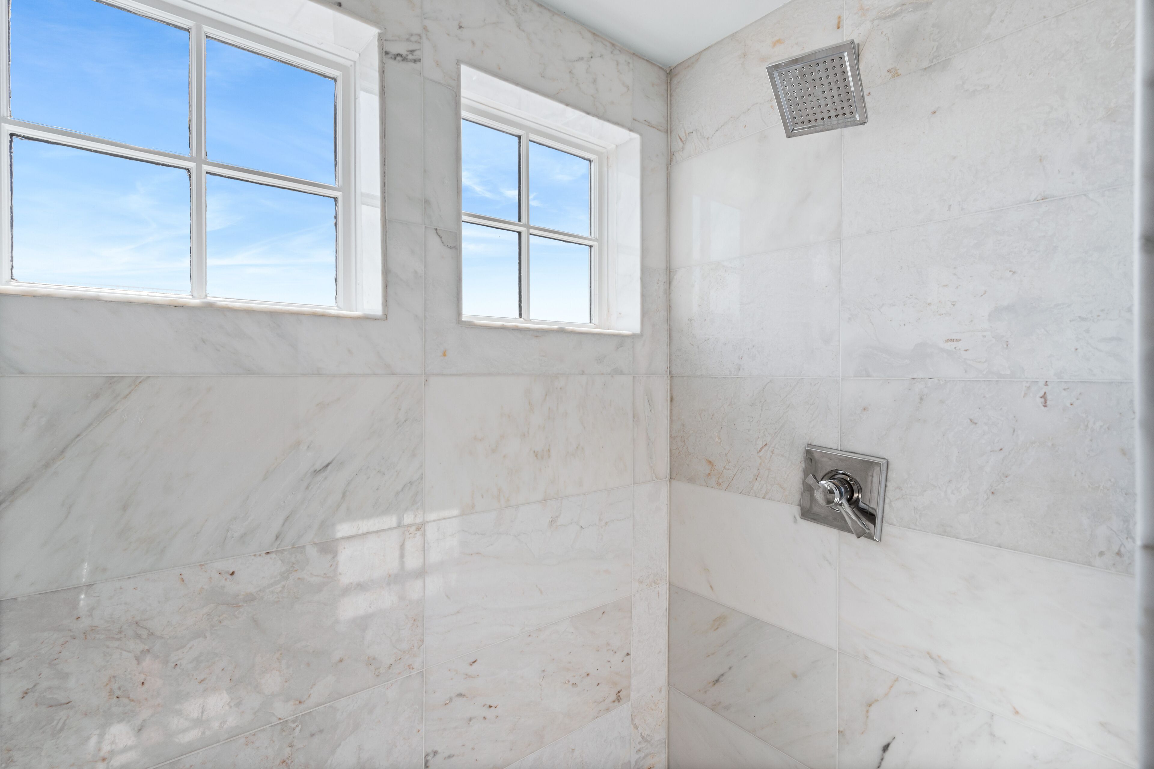 A bathroom with white marble walls, a shower with a rain showerhead, and two windows providing natural light.