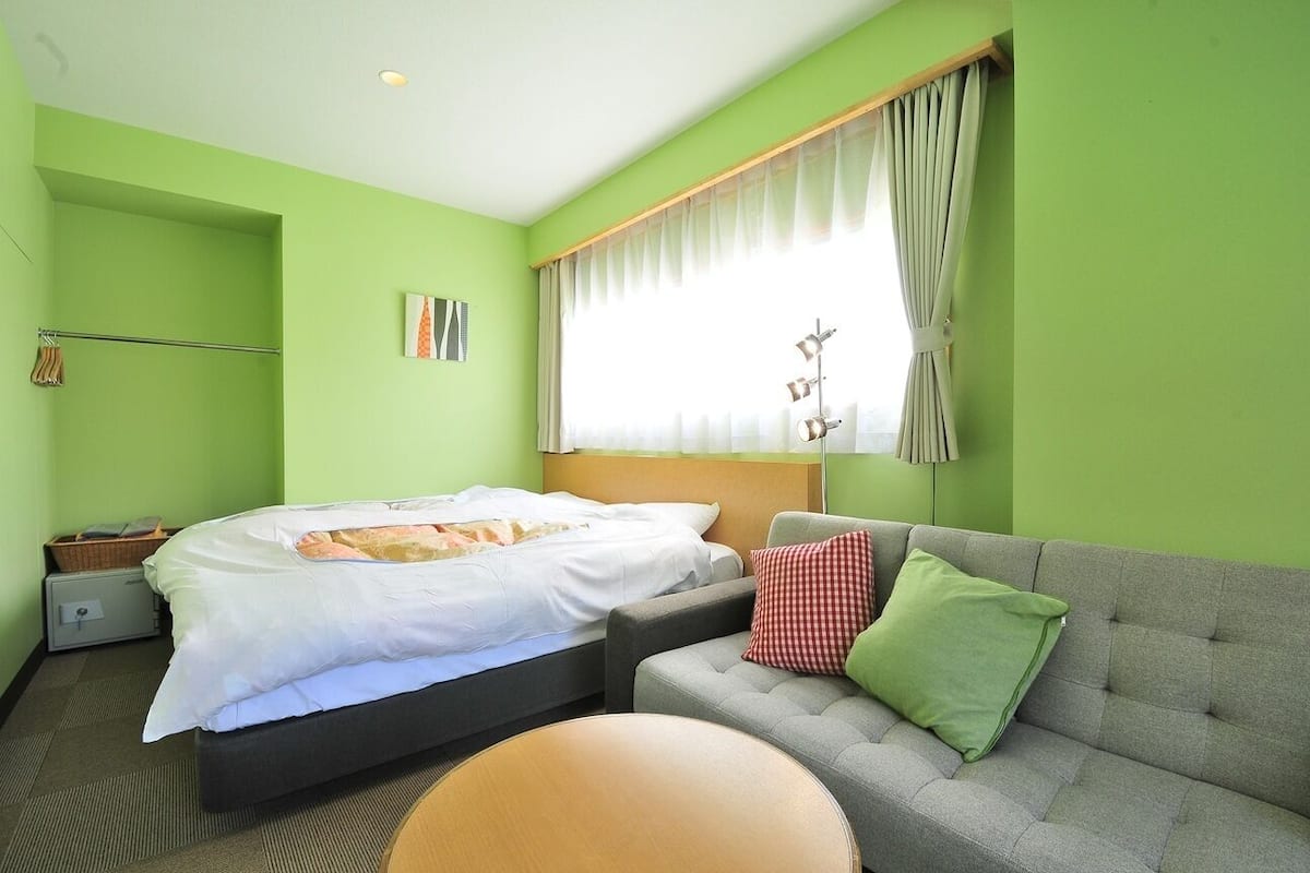 Quarto de hotel com cama, sofá cinza, mesa redonda pequena e janela com cortinas.