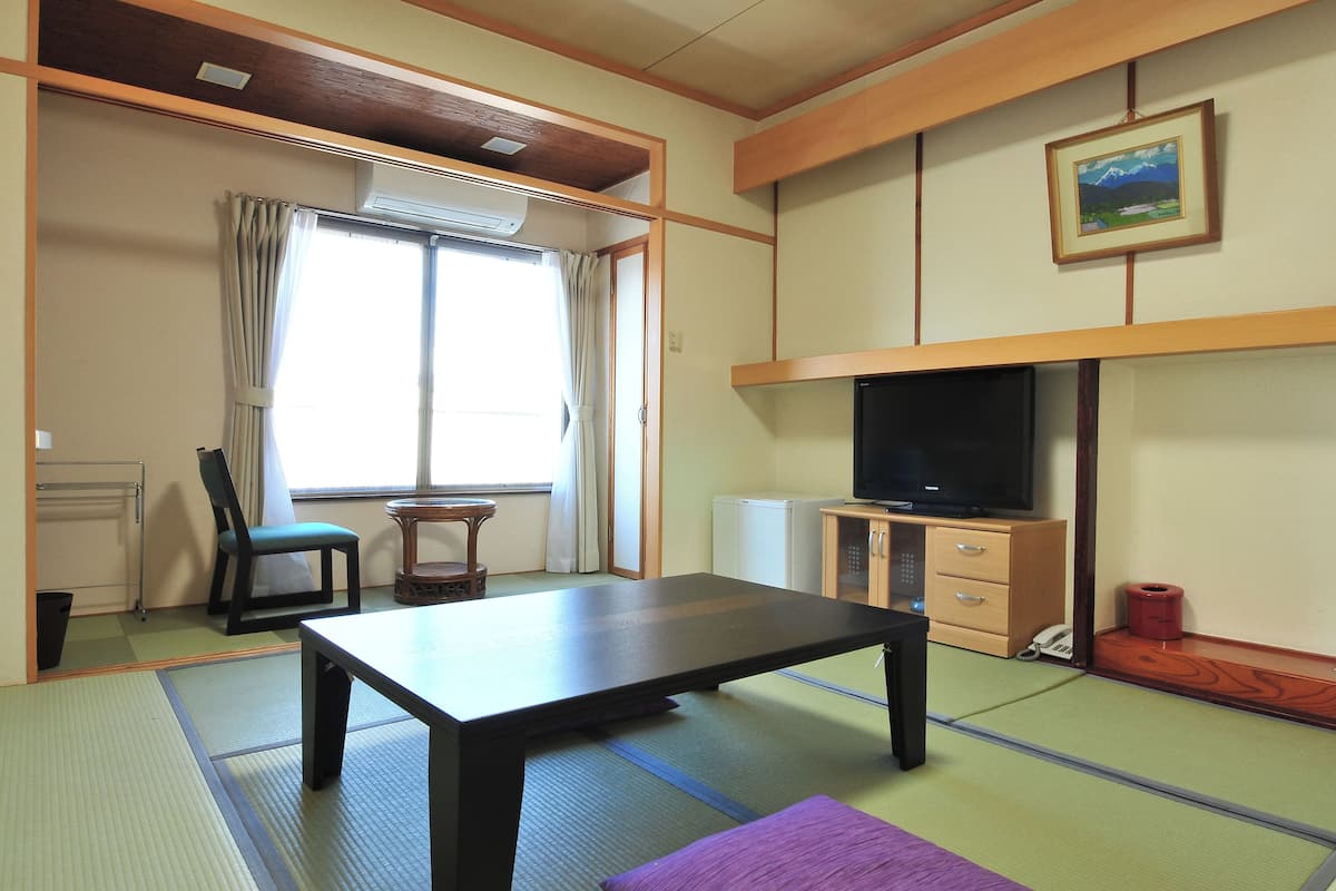 Um quarto tradicional japonês com piso de tatame, uma mesa baixa, uma cadeira, uma televisão e um pequeno armário.