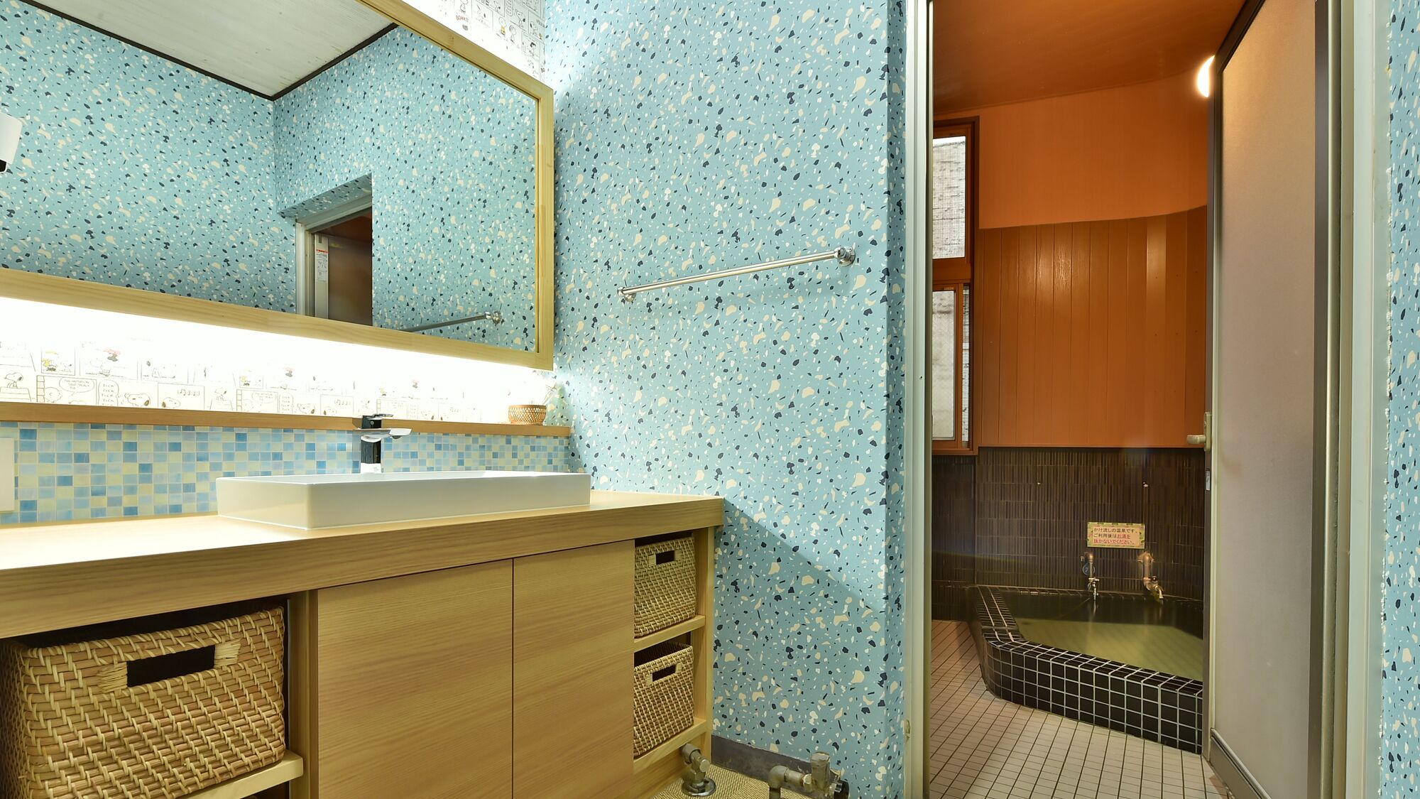 A bathroom with a blue speckled tile wall, a wooden vanity with a mirror, and a separate shower area with a black tiled wall.