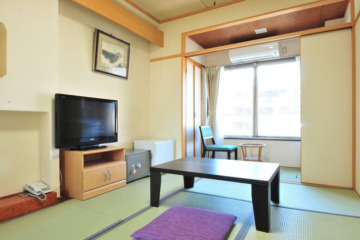 Quarto com piso de tatame, televisão sobre suporte de madeira, uma mesa pequena, um banco acolchoado e uma porta deslizante que dá para uma janela com cortinas.