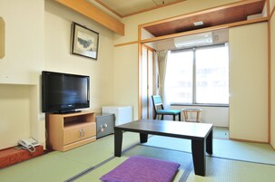 Una habitación con suelo de tatami, un televisor sobre un atril de madera, una mesita pequeña, un banco acolchado y un puertas corredera que da a una ventana con cortinas.