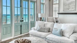 A spacious living room with a large window overlooking the sea, a white sofa with blue cushions, and a modern fireplace. - Beach Bungalow Cottage - Nestled behind the Dunes! Gulf Views! FREE GYM/TENNIS (Rosemary Beach)
