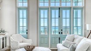 A room with a white sofa, a white armchair, and a round table with a decorative centerpiece. There are two large windows with a view of the ocean. - Beach Bungalow Cottage - Nestled behind the Dunes! Gulf Views! FREE GYM/TENNIS (Rosemary Beach)