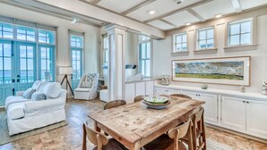 A spacious living area with a wooden dining table, white sofas, and large windows offering a view of the outdoors. - Beach Bungalow Cottage - Nestled behind the Dunes! Gulf Views! FREE GYM/TENNIS (Rosemary Beach)
