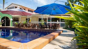 A swimming pool with tiled edges, surrounded by outdoor furniture and umbrellas, and a white fence. - Central Park Motel (Allenstown)