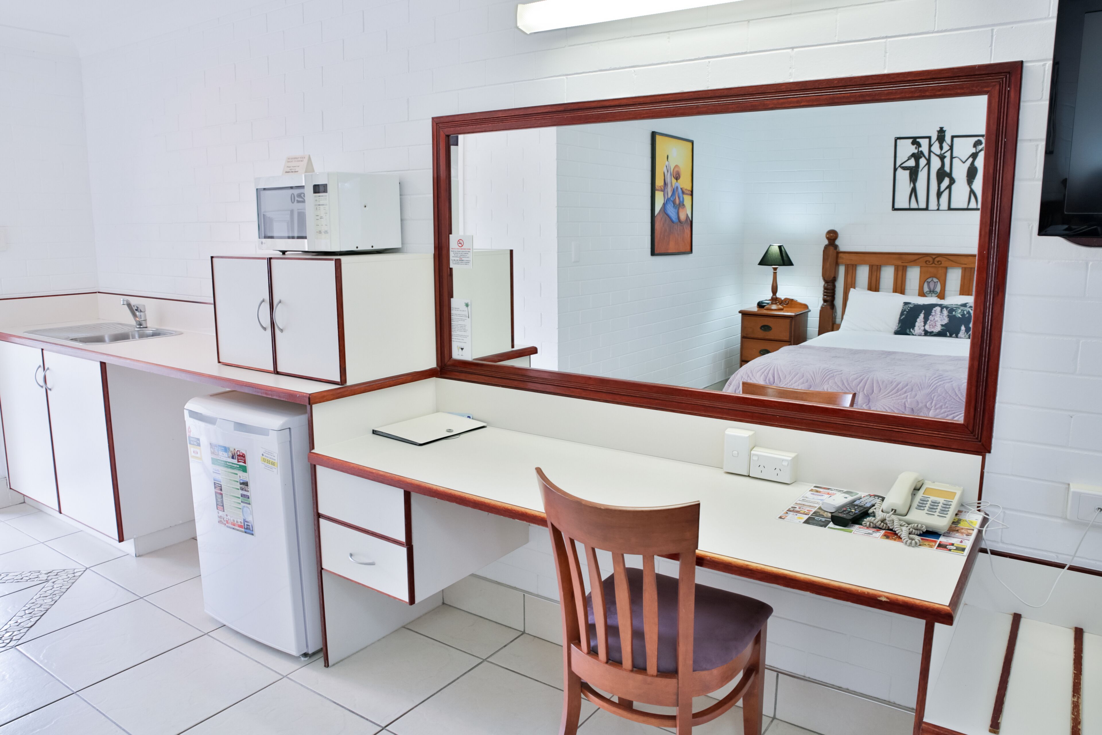 A compact hotel room with a kitchenette, a large mirror, a bed with a wooden headboard, a desk with a chair, and a television mounted on the wall.