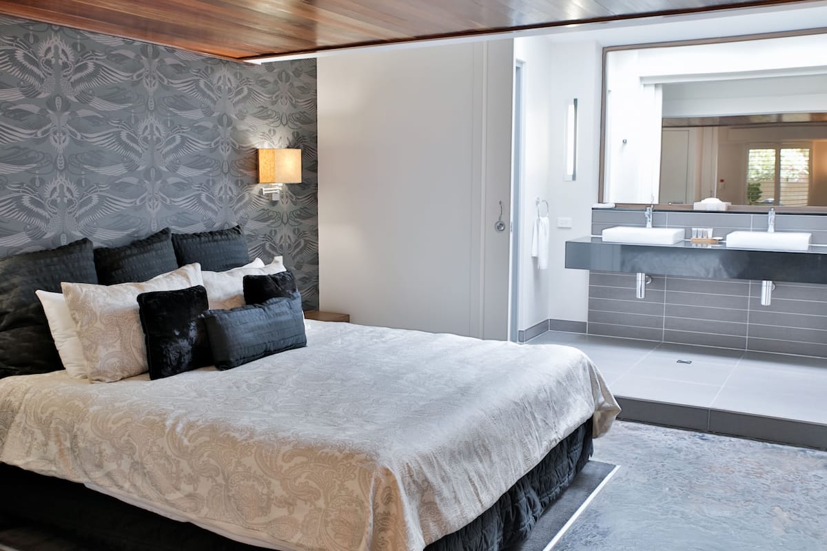 A modern bedroom with a large bed, a nightstand, a wall-mounted lamp, and a view of a bathroom with a sink and mirror.