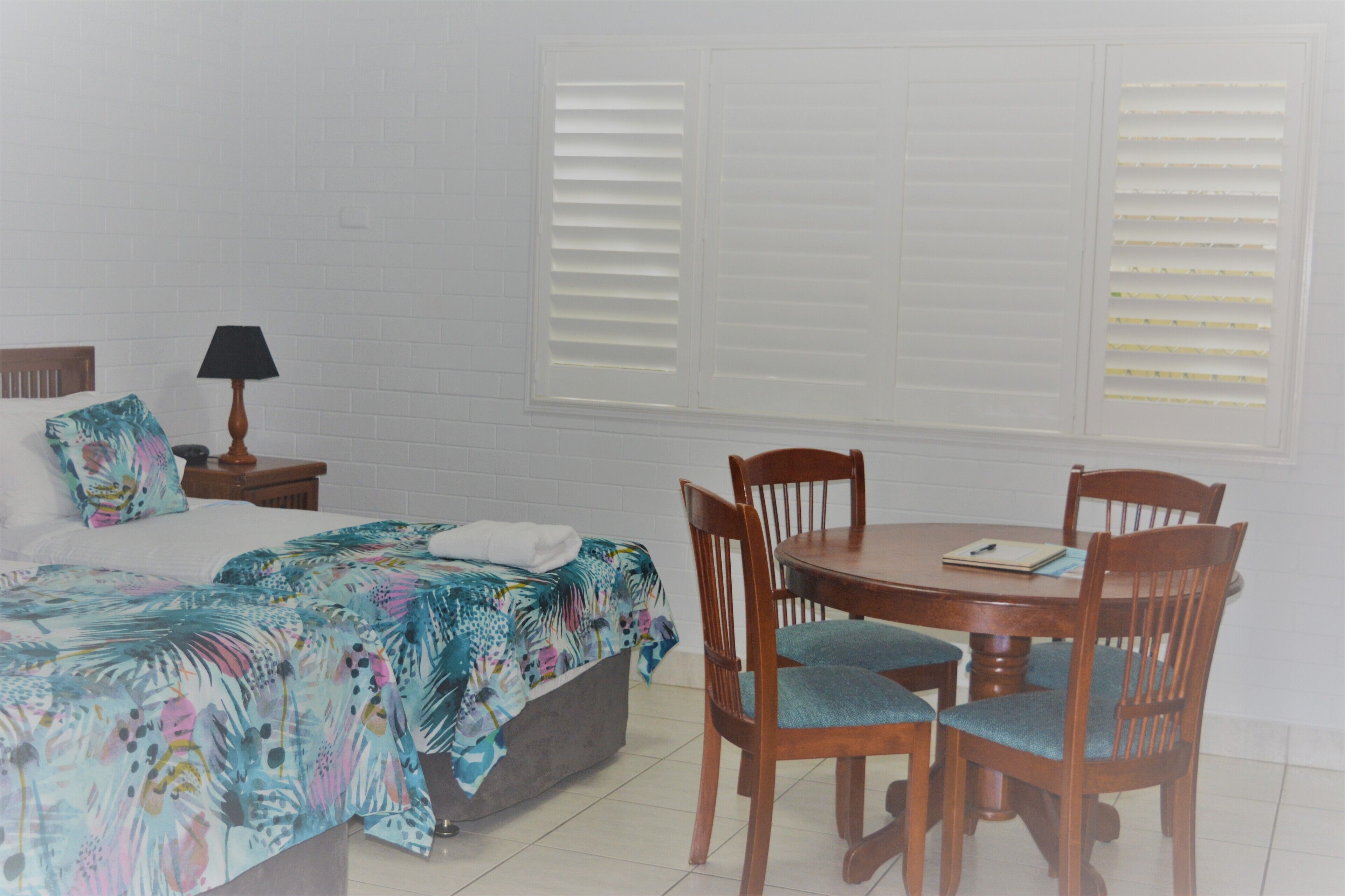 A bedroom with a bed, a bedside table with a lamp, a chair, a table, and a window with blinds.
