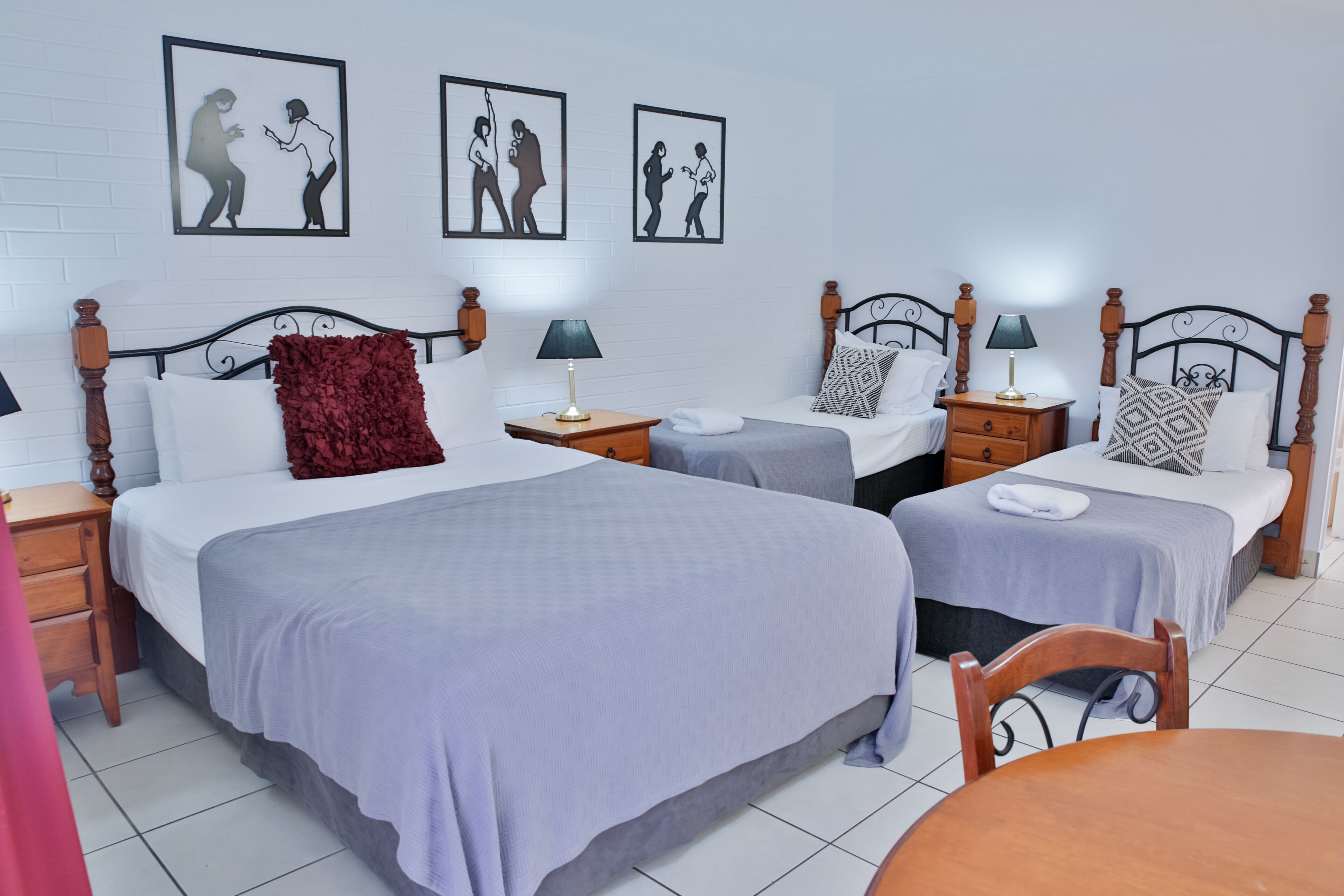 A hotel room with two single beds, a wooden nightstand with a lamp, a red throw pillow, and framed artwork on the wall.