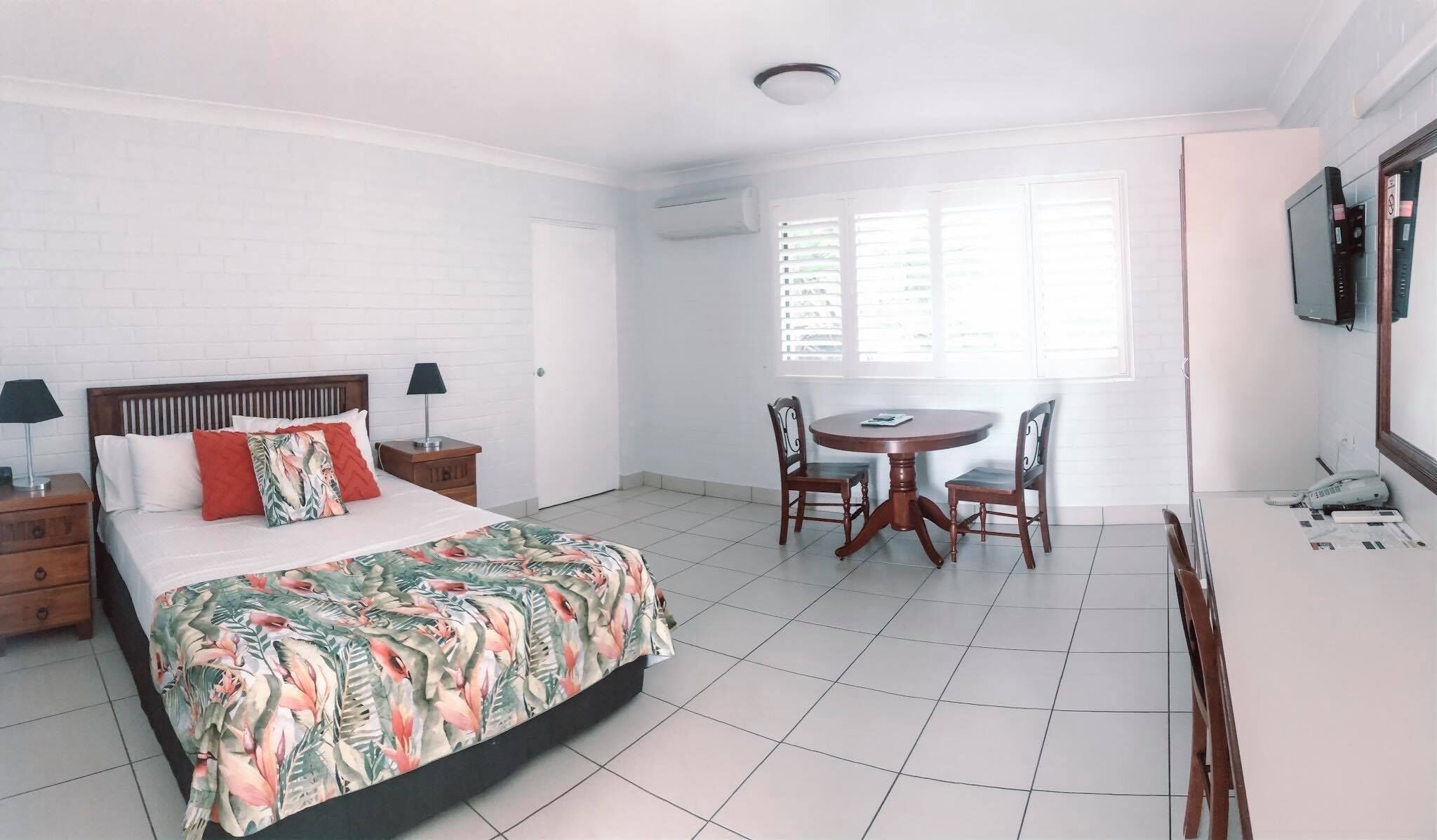 A hotel room with a bed, a desk, and a chair. There is a window with blinds, a TV mounted on the wall, and a telephone on the desk.