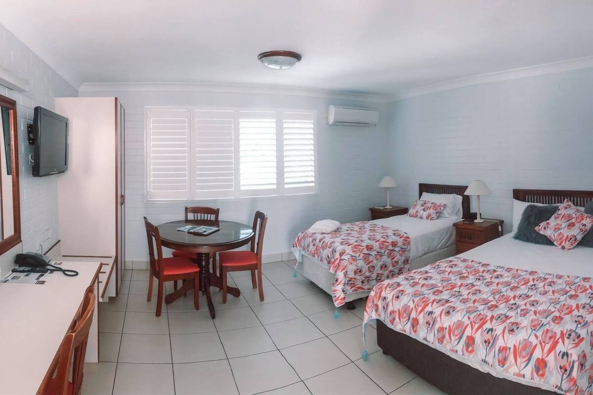 A hotel room with two beds, a dining table with chairs, a TV, and an air conditioning unit.