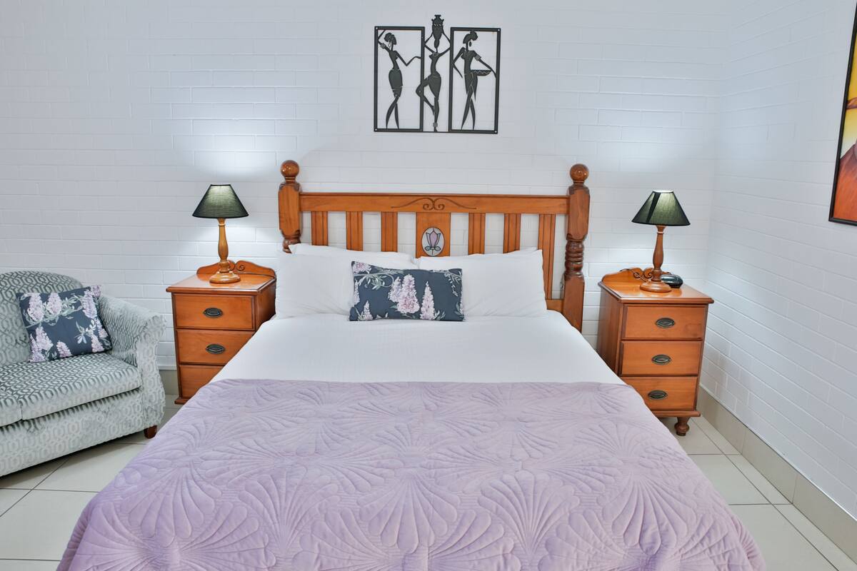 A bedroom with a large bed, two bedside tables, a wooden headboard, a chair, and a wall art piece.