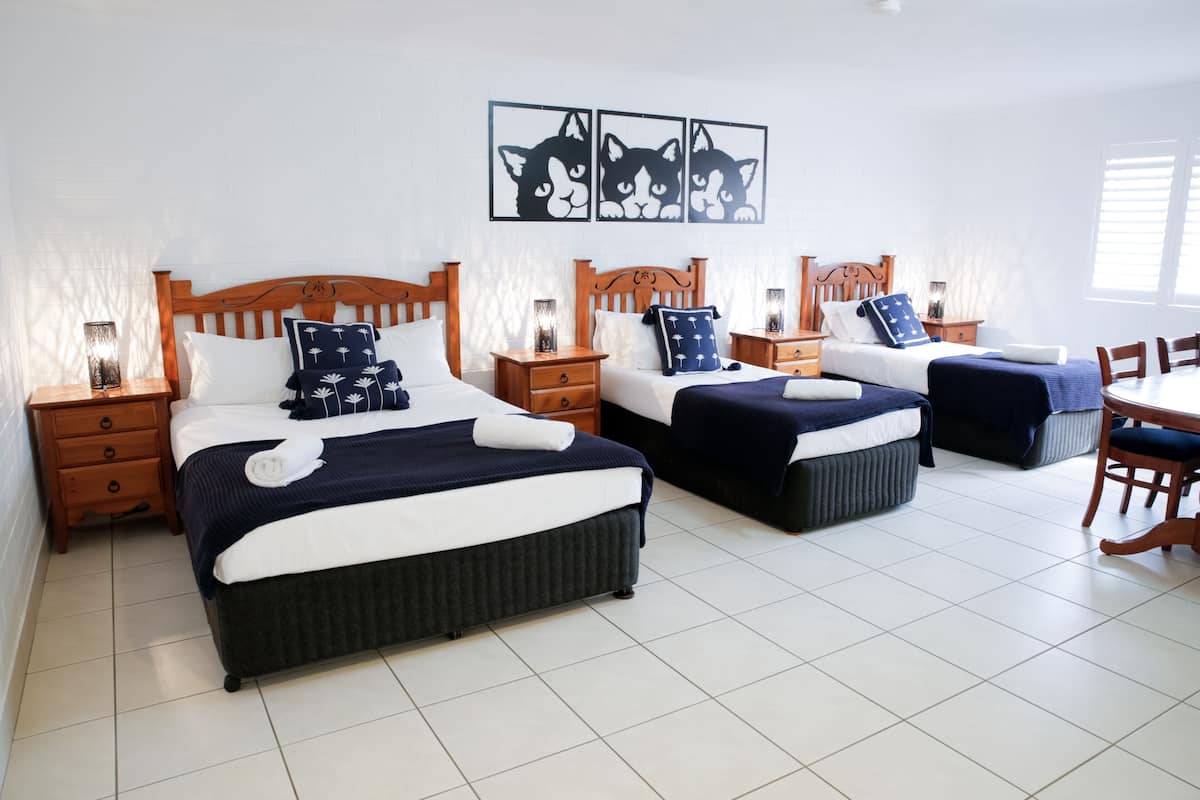 A hotel room with three beds, each with a wooden headboard, white bedding, and decorative pillows. There are bedside tables with lamps, a wall-mounted artwork of cats, and a dining area with a table and chairs.