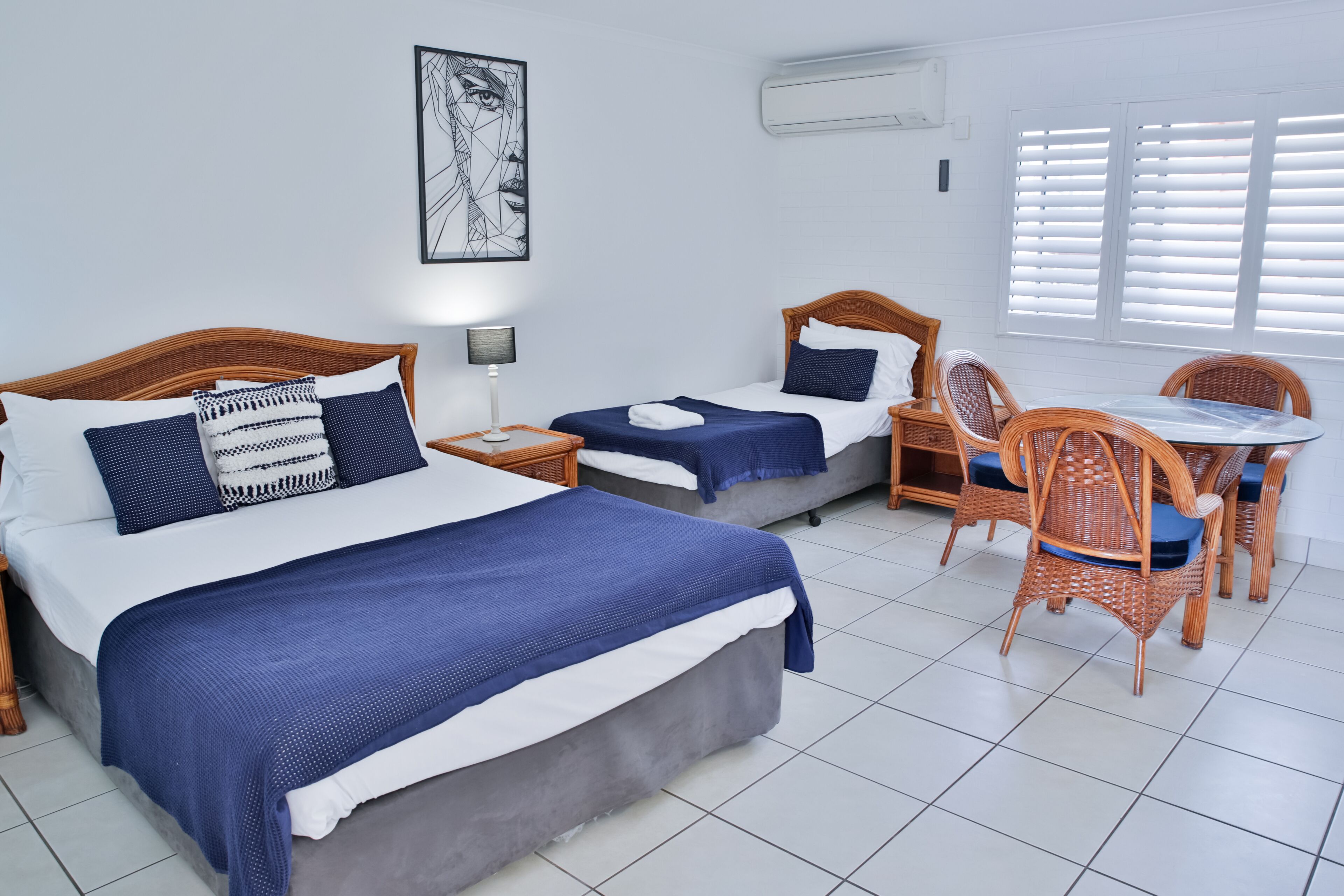 A hotel room with two beds, a dining table with chairs, a wall-mounted air conditioner, and a window with blinds.