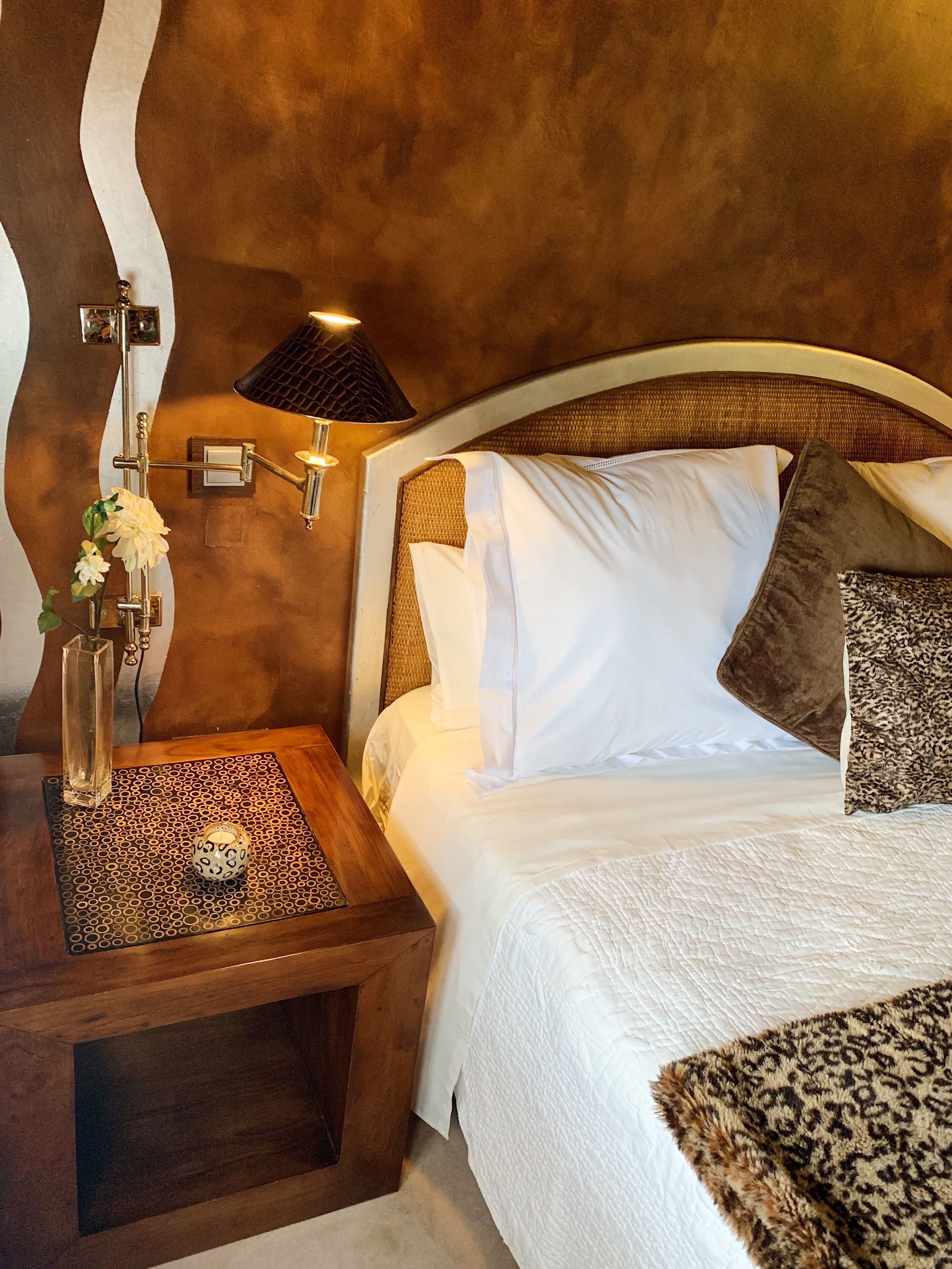 Una cama bien hecha con sábanas blancas y un plaid de leopardo, una mesita de noche con un jarrón de flores, y una lámpara fijada a la pared.