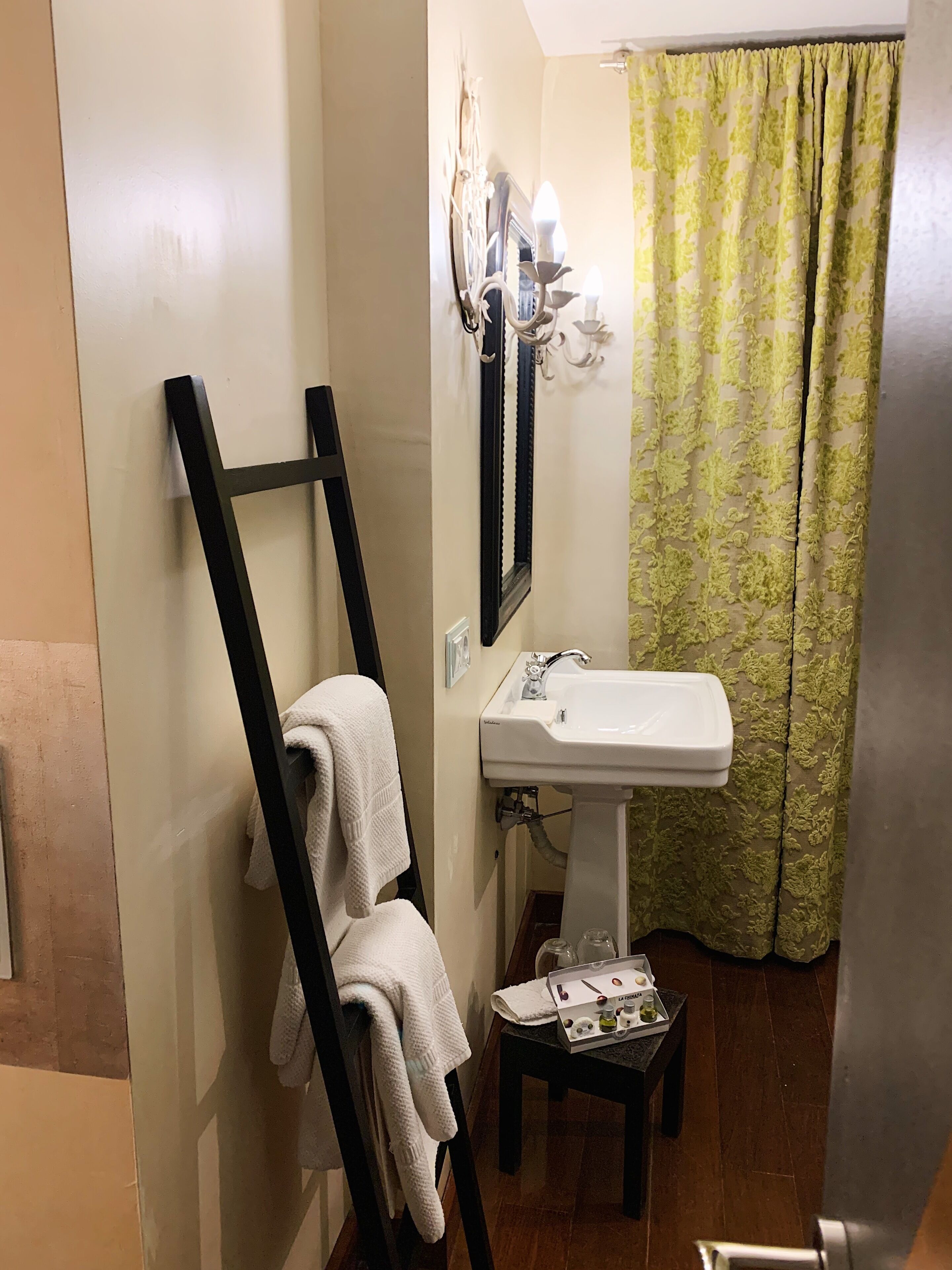 Un baño con lavabo blanco, un perchero con toalla, una mesita con artículos de aseo y una cortina de ducha verde con estampado.