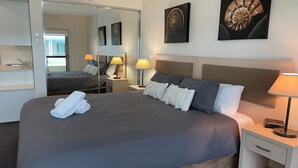 A hotel room with a large bed, a bedside table with a lamp, a mirror, and artwork on the wall. - Pacific Blue  245-265 Sandy Point Rd - Dual Key Access, air conditioned unit with resort facilities and linen supplied (Salamander Bay)