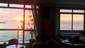 A living room with a large window overlooking the sea at sunset, a sofa, a small table with a remote control, and a potted plant.
