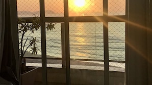 A room with a view of the sunset over the ocean, a window with a metal grill, and a potted plant on the balcony.
