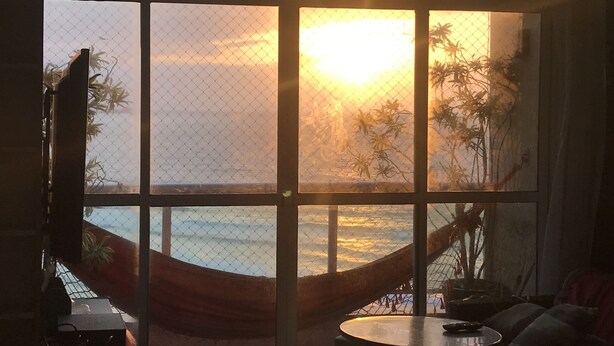 A room with a large window overlooking a beach at sunset.