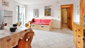Een lichte woonkamer met een rode bank, een houten tafel met een tafelkleed met hartenprint, een keuken met houten kastjes, en een groot raam dat natuurlijk licht binnenlaat.