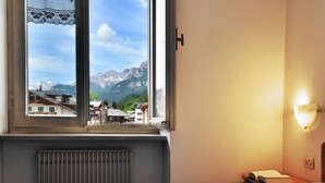 Hotelkamer met een bed, een radiator, een raam met uitzicht op bergen en gebouwen, en een wandlamp.