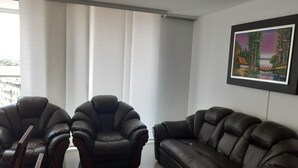 Ein modernes Wohnzimmer mit zwei schwarzen Ledersofas und einem Sessel, einem großen Fenster mit weißen Vorhängen und einem gerahmten Gemälde an der Wand.