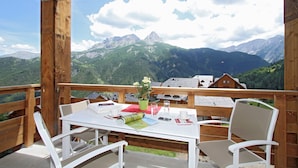 A wooden deck with a table and chairs, overlooking a mountainous landscape with houses. - Studio avec Wi-Fi Gratuit + Accès Piscine! (Pra Loup)
