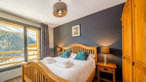 A hotel room with a wooden bed, a nightstand, a lamp, a window with a view of a mountain, and a framed picture on the wall. - Flat 6 pers - modern, central, ski in ski out (CHATEL)