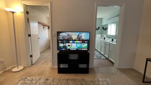 Una sala de estar con un televisor de pantalla plana sobre un mueble bajo, piso alfombrado y un pasillo que da a una cocina.