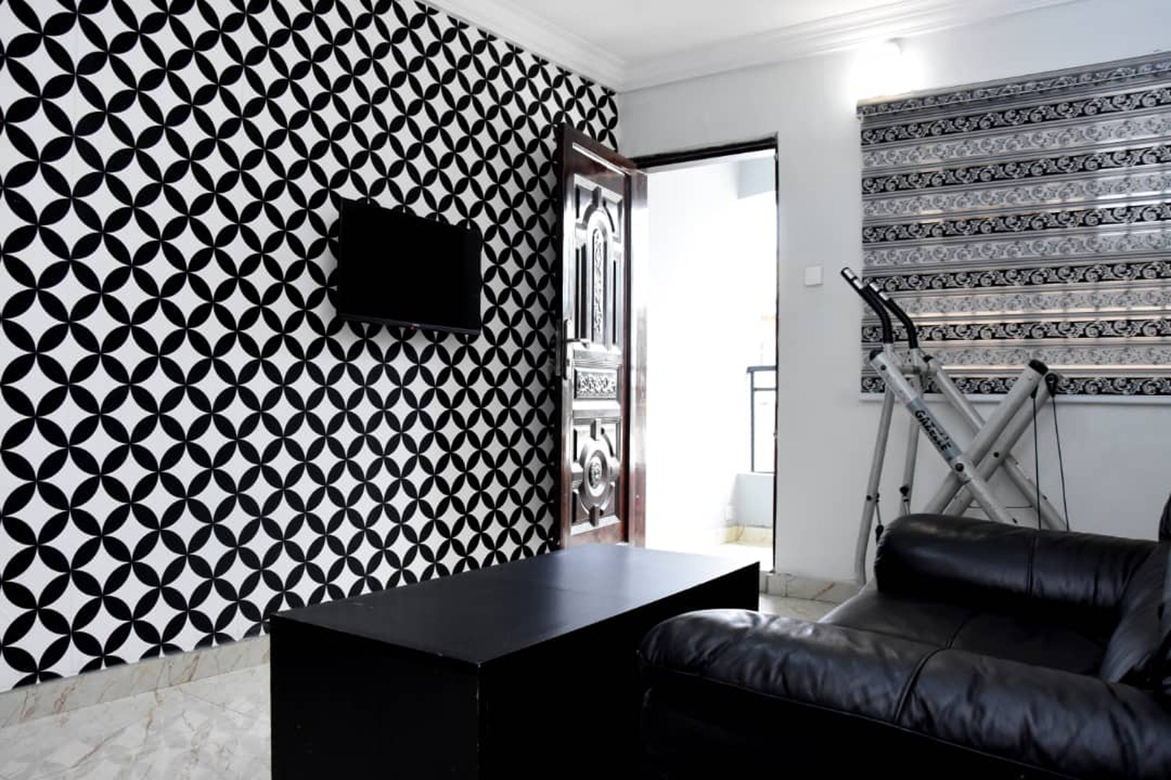 A room with a geometric patterned wall, a black leather sofa, a flat-screen TV mounted on the wall, and a window with a view of the outside.