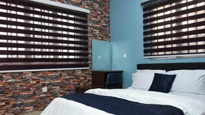 A bedroom with a bed, a desk, and a chair. The room has a stone wall and a window with blinds. - PrimeMeridian 24/7 Electricity Hotel & Restaurant (Lekki)