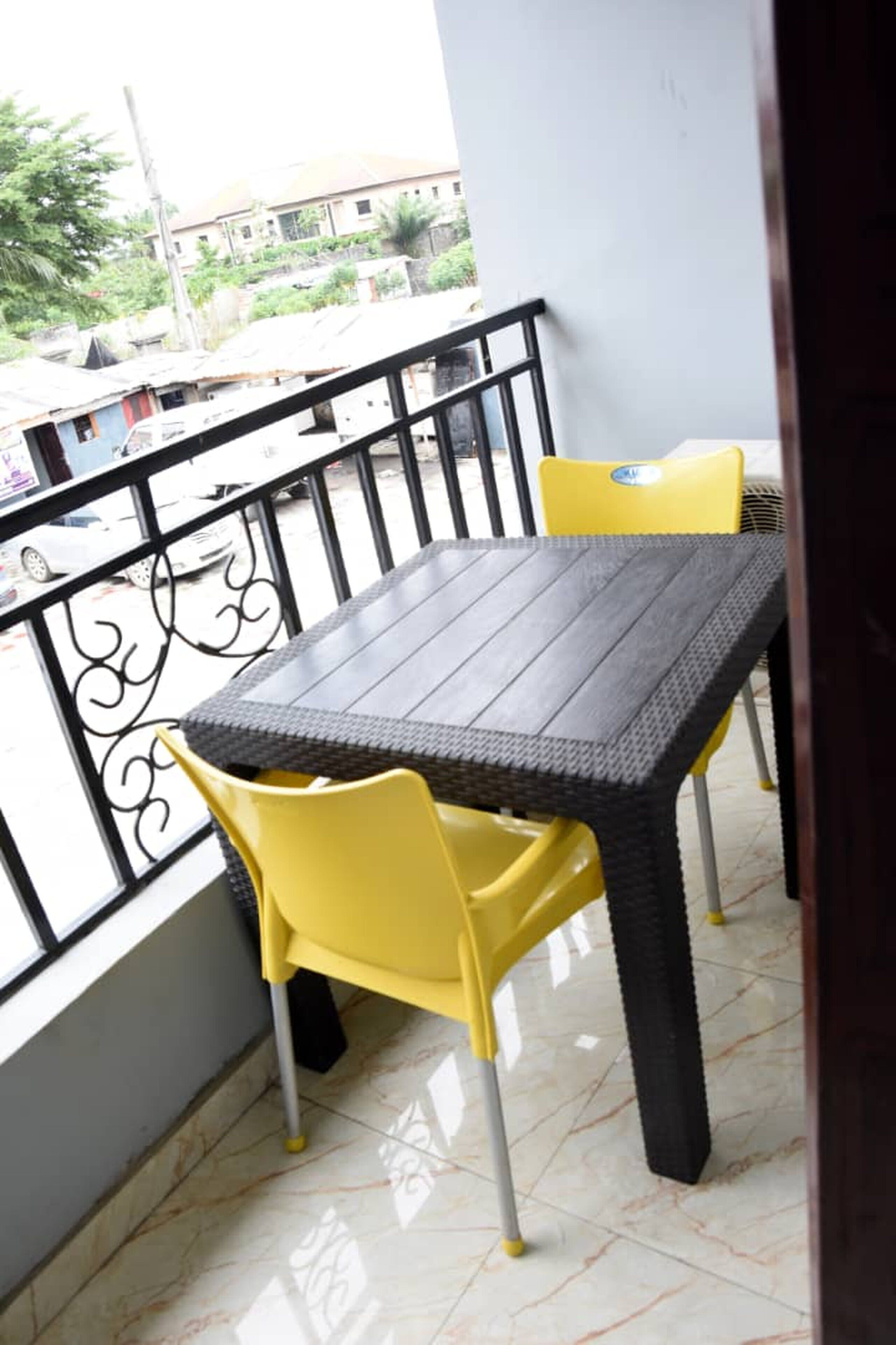 Un balcon avec une table et des chaises jaunes, un sol carrelé et une vue sur des bâtiments et des véhicules en arrière-plan.