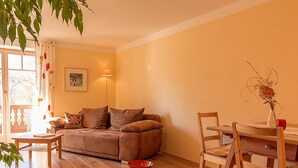 Een gezellige woonkamer met een bruine bank, een houten eettafel en stoelen. Er staat een rode schaal op de vloer en een vaas met gedroogde bloemen op de tafel.