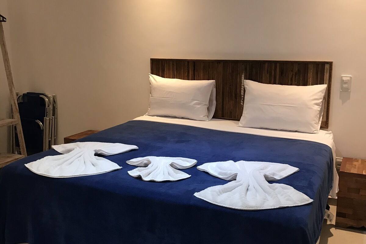 Ein ordentlich bezogenes Bett mit blauer Bettdecke und weißen Kissen, ein Holzkopfstück und ein Nachttisch mit Lampe.