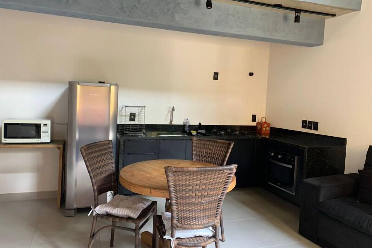Eine kompakte Küche mit Essbereich, darunter ein runder Holztisch und Korbstühle, eine Mikrowelle, ein Kühlschrank sowie eine schwarze Arbeitsplatte mit Spüle.