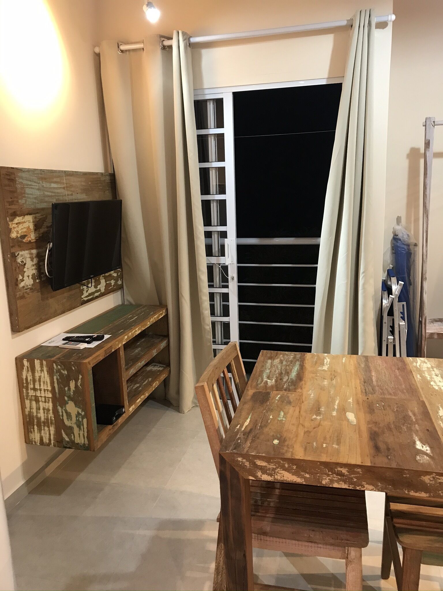 A room with a wooden dining table and chairs, a television mounted on a wooden shelf, and a window with curtains.