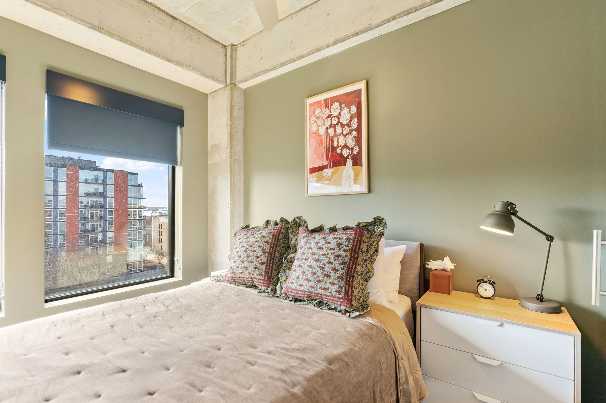 A bedroom with a large bed, a nightstand, a lamp, a framed picture, and a window with a city view.
