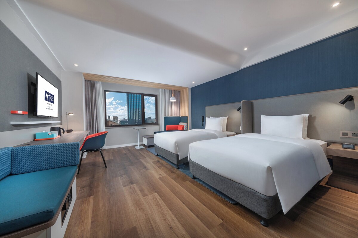 A modern hotel room with two beds, a sofa, a desk, and a TV.