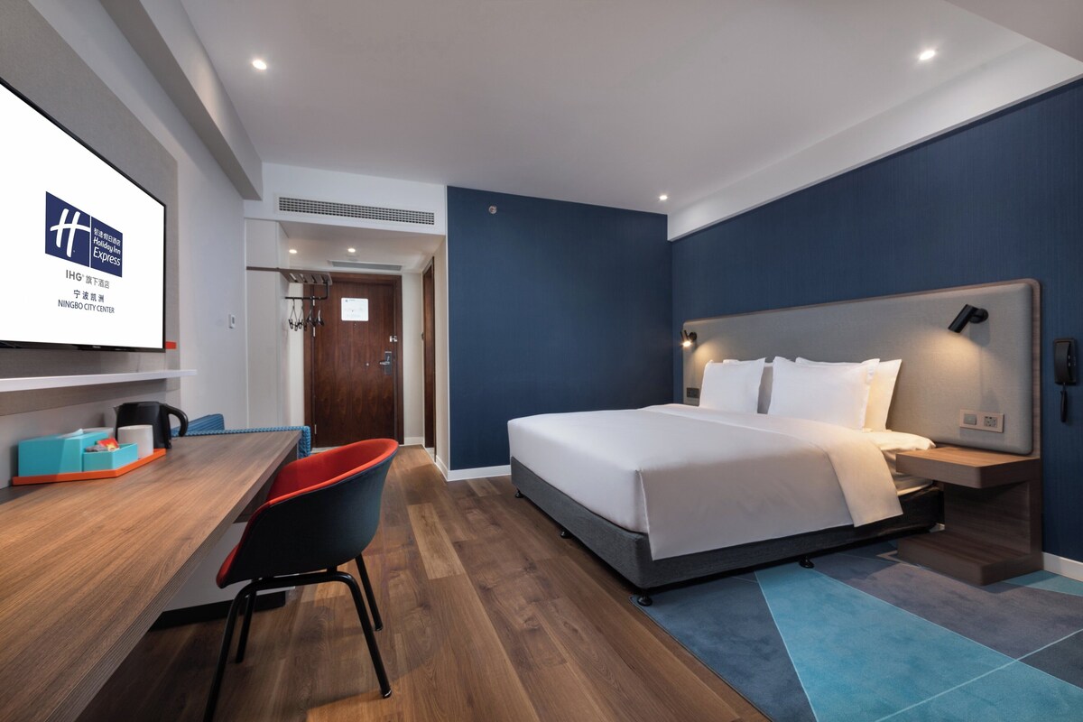 A modern hotel room with a large bed, a desk with a chair, and a TV screen displaying the hotel name.