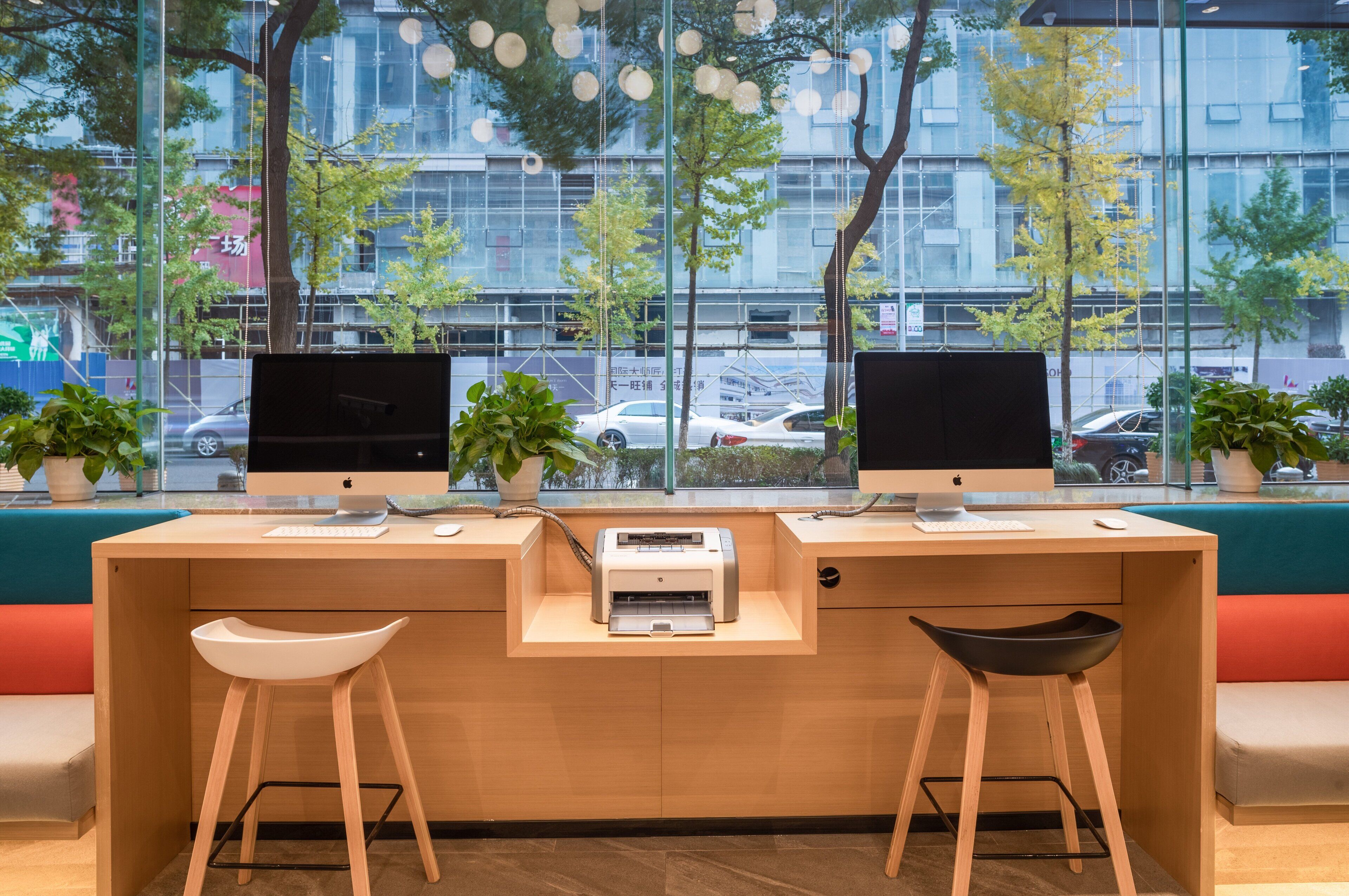 A modern office space with two desks, each with a computer monitor, keyboard, and mouse. There are two chairs, one white and one black, and a printer on the desks. The room has large windows with a view of a city street and trees.