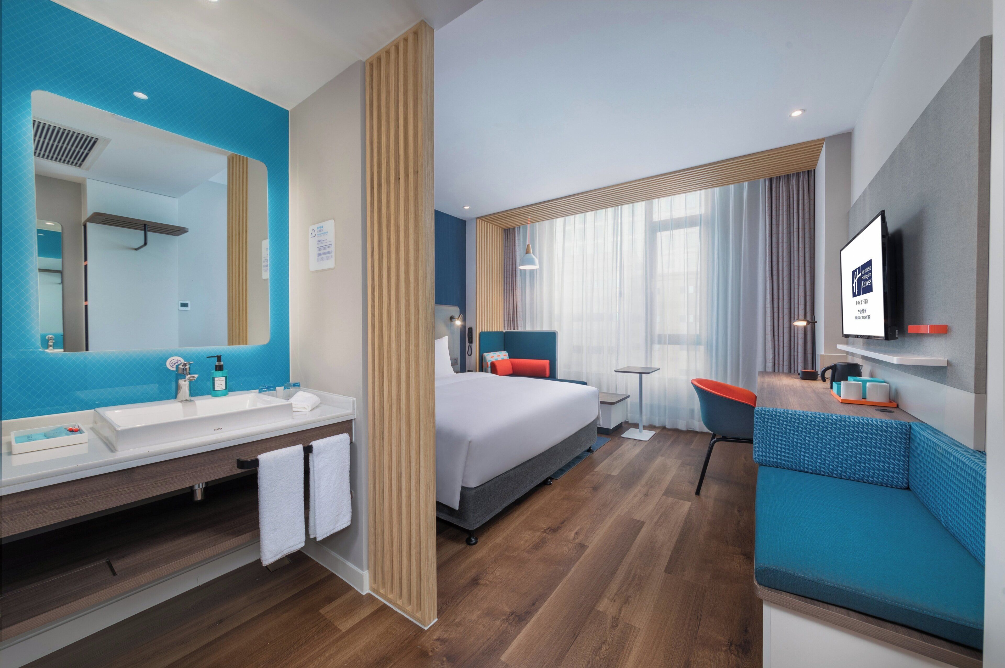 A modern hotel room with a large bed, a bathroom with a sink and mirror, a small table, and a TV mounted on the wall.