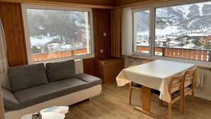 Et hyggeligt værelse med en sofa, et lille bord og et spisebord med stole. Store vinduer giver udsigt over et sneklædt landskab.