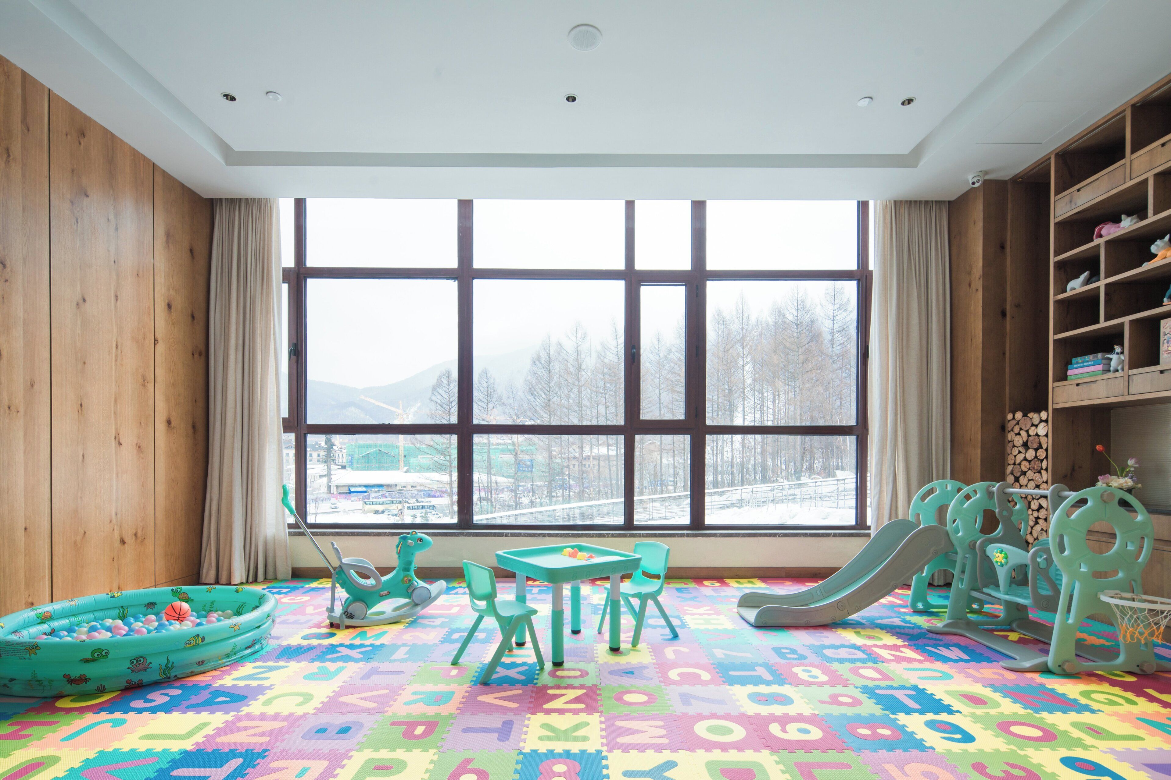 A children's playroom with a large window, colorful play mat, and various toys.