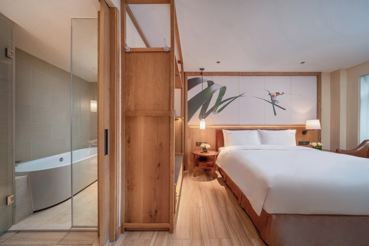 A hotel room with a large bed, a wooden wardrobe, a small bedside table with a lamp, a wall-mounted painting, and a shower area with a glass door.