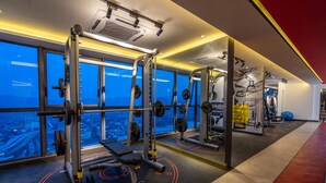 Un gimnasio con equipo de levantamiento de pesas, ventanas amplias y vista a la ciudad.