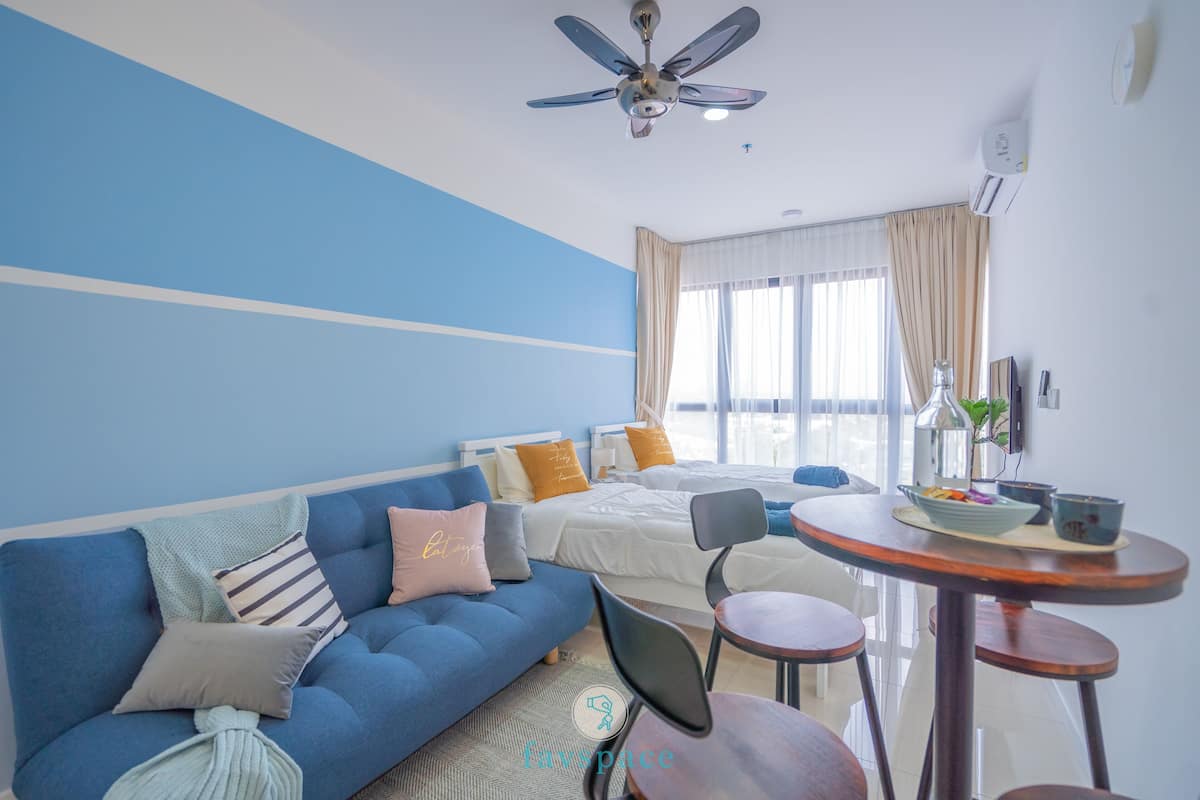 A modern hotel room with a blue accent wall, a bed with white bedding, a blue sofa with multiple pillows, a round wooden dining table with chairs, and a ceiling fan.